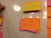 A close up of large yellow, orange, and pink sticky notes stuck in two piles to a whiteboard, with other sticky notes at the edges of the photo. The note at the top of the bottom pile says "How to Study/Approach them" and the note at the tops just says "EPISTEMICS"